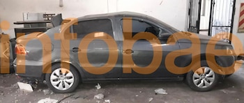 Un sedán oscuro, posiblemente un Volkswagen, se ve de perfil estacionado en un garaje con piso de cemento y objetos diversos alrededor