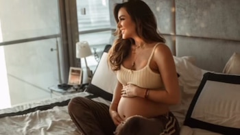 Mujer embarazada sentada en una cama, con top de tirantes y pantalón, mirando hacia la ventana. Toca su vientre. Hay una lámpara de mesa y una ventana detrás
