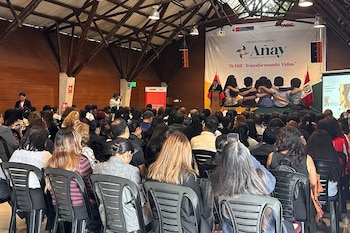 Vista trasera de una audiencia sentada en sillas de plástico negras, mirando un escenario con un podio, dos banderas peruanas y un banner del Primer Encuentro Añay
