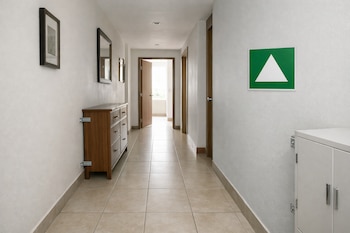 Pasillo de vivienda con paredes blancas y suelo de baldosas; cómoda de madera y armario blanco anclados a la pared; cartel verde de zona segura y cuadros.
