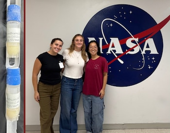 Tres mujeres de pie delante del logotipo de la NASA y, a la izquierda, dos tubos de laboratorio de plástico con tapas azules