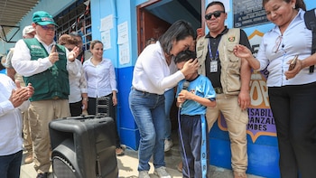 Mujer de camisa blanca besa la frente de niño con uniforme escolar y micrófono. Hombres y mujeres adultos observan. Hombre con chaleco verde. Altavoz negro