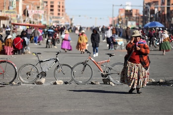 Una mujer con sombrero y vestimenta tradicional andina camina en una calle bloqueada por dos bicicletas y rocas en La Paz, con más personas al fondo