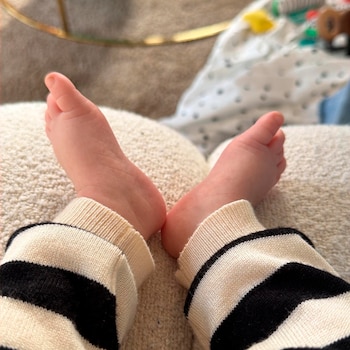 Primer plano de los pies rosados de un bebé, calzados con un pantalón o pijama de rayas horizontales blancas y negras, descansando sobre una superficie clara y texturizada