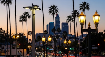 Paisaje urbano de Los Ángeles al anochecer, con farolas encendidas, palmeras altas, edificios de silueta y estelas de luces de vehículos en las calles.