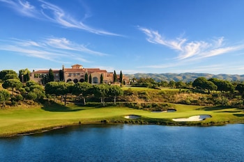 Un campo de golf con césped verde, trampas de arena, árboles, un lago en primer plano y una gran edificación frente a montañas