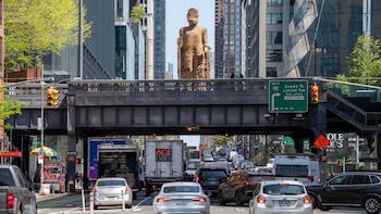 Réplica del Buda de Bamiyán, una estatua alta de color arena, se asoma sobre un puente elevado de metal en una calle urbana concurrida, rodeada de edificios