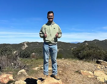 Un hombre joven con gafas, sudadera verde, jeans y zapatillas deportivas, de pie sobre una roca, hace el gesto de pulgares arriba en un paisaje montañoso bajo un cielo azul