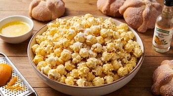 Tazón de palomitas de maíz con azúcar, acompañado por panes de muerto, un rallador con cáscara de naranja, mantequilla y una botella de azahar.