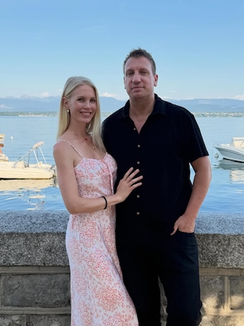 Un hombre y una mujer posan junto a un lago con montañas al fondo. El hombre usa camisa negra, la mujer un vestido floral claro. Hay botes en el agua