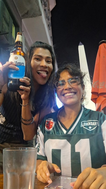 Dos mujeres sonriendo. Una sostiene una botella de cerveza. La otra lleva anteojos y camiseta. Un vaso y teléfono móvil en la mesa. Obelisco en segundo plano