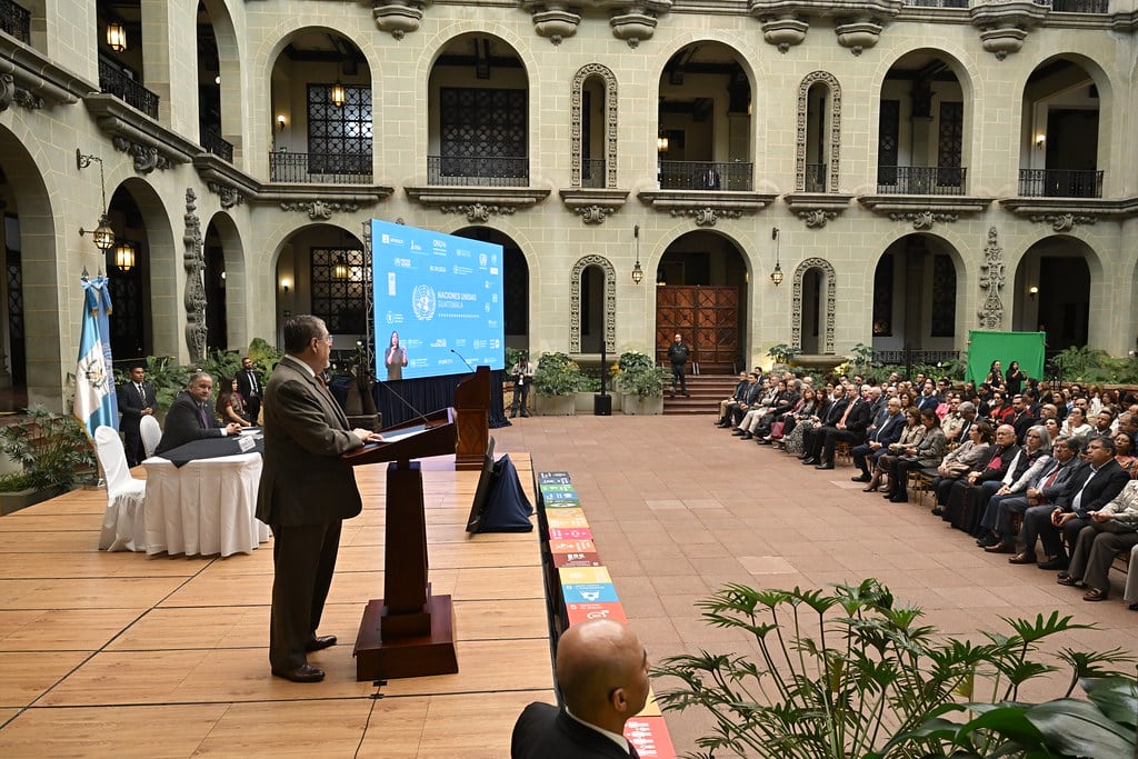Presidente Bernardo Arévalo se dirige a una audiencia en una convención de Naciones Unidas, celebrada en un patio interior con una impresionante fachada arquitectónica y una gran pantalla mostrando el evento. (Secretaría de Comunicación Social de la Presidencia de Guatemala)