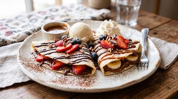 Plato con crepas dobladas rellenas de fruta, acompañadas de helado, nata montada, fresas, arándanos y jarabe sobre una mesa de madera.
