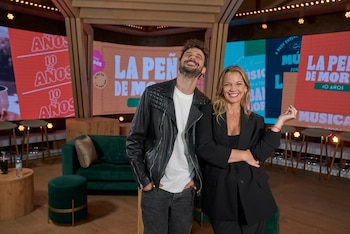 Diego Leuco y Carina Zampini de pie y sonriendo en un set de televisión. Él lleva chaqueta de cuero negra, ella un blazer negro. Pantallas LED de fondo