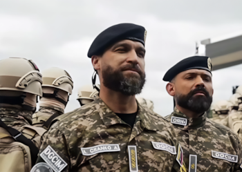 Primer plano de dos hombres con barba, boinas negras y uniforme militar camuflado. Detrás hay otros militares con cascos y uniformes similares