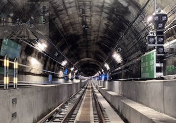 Interior de un túnel con vías de tren, luces en las paredes, señal luminosa, señales verdes y barreras amarillas y negras
