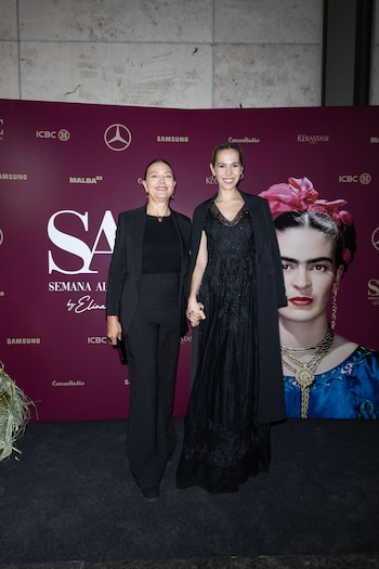 Dos mujeres vestidas de negro posan sujetándose de la mano ante un mural publicitario que incluye un retrato de Frida Kahlo