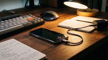 Primer plano de un celular conectado a su cargador, con la pantalla hacia abajo, sobre un escritorio de madera, junto a un teclado, ratón y libreta.