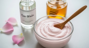 Un tazón de vidrio con mascarilla de arcilla rosa cremosa y una cuchara de madera, junto a una botella de agua de rosas y un frasco de miel con pétalos rosas.