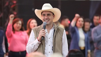 Hombre con sombrero vaquero blanco, camisa a cuadros y chaleco beige, sonriendo y sosteniendo un micrófono, con personas difusas en el fondo