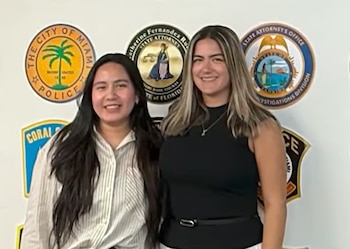 Dos mujeres posan juntas sonriendo frente a una pared blanca decorada con sellos y emblemas policiales oficiales