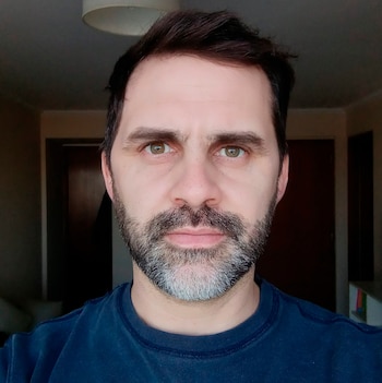 Hombre con cabello oscuro y barba canosa mira de frente. Viste un suéter azul. Se observa una lámpara de techo y una puerta al fondo