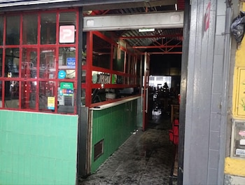 Entrada de un local comercial con puerta gris entreabierta. Marcos rojos en ventanas con calcomanías de pago. Azulejos verdes y un interior oscuro con mesas