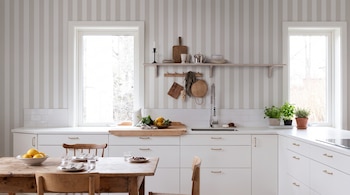 Cocina con pared de rayas, dos ventanas, armarios blancos, encimera, fregadero, estante con utensilios, hierbas en maceta, mesa de madera, sillas, limones.