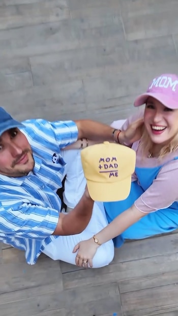 Jorge Blanco y una mujer rubia sonríen sentados en un suelo de madera. Él sostiene una gorra amarilla que dice 'MOM + DAD = ME'