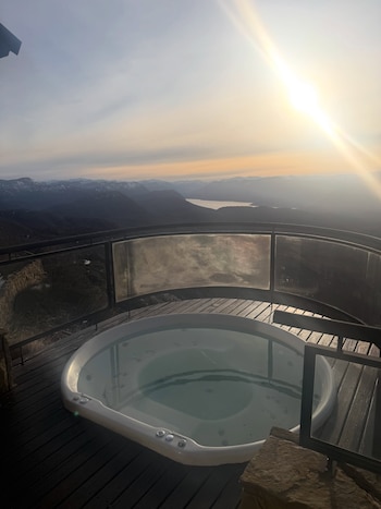 Un hidromasaje circular en una terraza de madera con barandilla de vidrio frente a un paisaje de montañas y un lago bajo la luz del sol