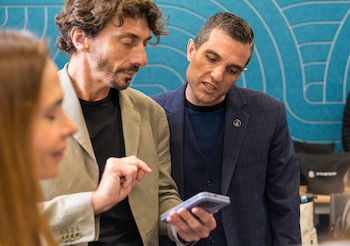 Ramiro Sánchez, de barba y chaqueta caqui, señala un smartphone que sostiene, mientras Leandro Petersen, en traje azul con pin de AFA, lo observa. Una mujer en primer plano, desenfocada, a la izquierda