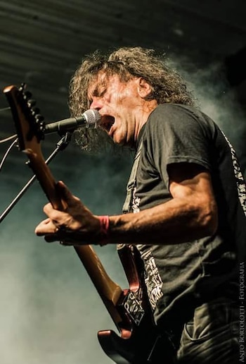 Un hombre de pelo largo rizado canta en un micrófono y toca una guitarra eléctrica. Lleva una camiseta oscura y una pulsera roja, rodeado de humo