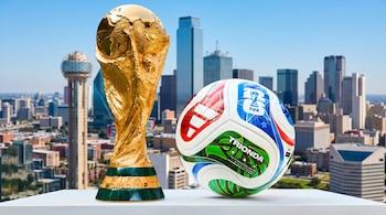 Copa Mundial FIFA dorada y balón oficial Trionda en primer plano. Detrás, el skyline de Dallas, Texas, bajo un cielo azul. Los objetos están sobre una superficie blanca.