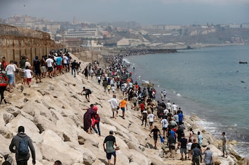 Una multitud de personas camina por una costa rocosa, junto al mar, una valla de alambre de espino y edificaciones en la distancia
