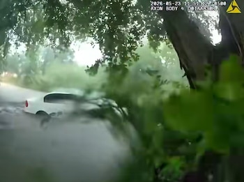 Un coche blanco parcialmente sumergido y arrastrado por una corriente de agua turbia, rodeado de vegetación y árboles verdes, bajo un cielo brillante y nublado