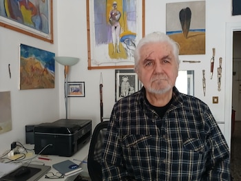 Retrato de Enrique Breccia, un hombre de cabello y barba blancos, vistiendo una camisa a cuadros, en una habitación con varias pinturas y un escritorio con impresora