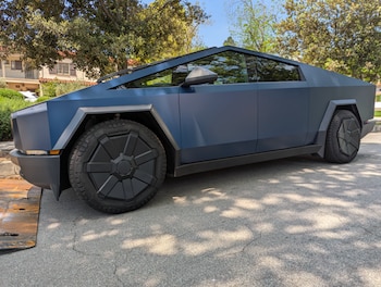 Un Tesla Cybertruck de color oscuro, posiblemente azul mate o negro, estacionado en una calle de asfalto con árboles y casas al fondo