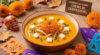 Tazón con crema de cempasúchil, cubos de queso, adorno floral, calavera decorativa y papel picado sobre una mesa de madera.