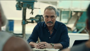 Salvador del Solar con bigote y camisa azul oscura sentado ante una mesa con un cuaderno, observando a dos personas vistas de espaldas