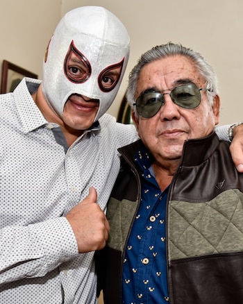 Un hombre con máscara blanca y camiseta estampada, con el pulgar arriba, abraza a un hombre mayor con gafas de sol, camisa azul y chaqueta acolchada