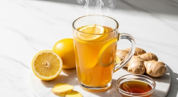 Primer plano de una taza de cristal con té de jengibre, limón y miel humeante, sobre mármol blanco, con rodajas de limón, jengibre fresco y un cuenco de miel.