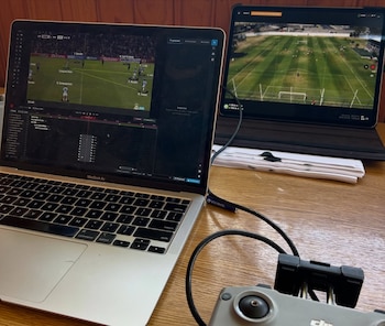 Una MacBook Air con software de análisis de fútbol y un iPad mostrando un campo de juego, conectados a un controlador de dron sobre una mesa de madera