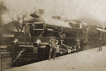 Locomotora de vapor negra número 38 con la palabra "TUCUMAN" y cuatro hombres con ropa de época posando en una plataforma de estación