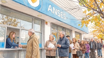Fila de personas mayores ante una oficina de ANSES donde un hombre saluda a una empleada en la ventanilla.