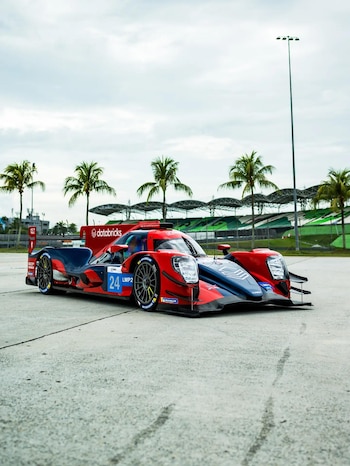 Un automóvil de carreras rojo y azul con el número 24 sobre una pista de concreto frente a palmeras y tribunas