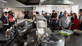 Varias personas, incluyendo actores y equipo técnico, se encuentran en una cocina industrial con cámaras, trípodes, ollas, platos y vegetales sobre mesadas