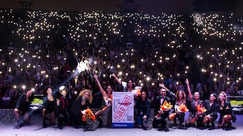 Doce artistas posan en un escenario, sostienen flores y un cartel de Ticketmaster con “SOLD OUT”, frente a una audiencia iluminada por luces de celulares