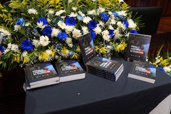 Varios libros titulados 'Maras y pandillas en Guatemala' sobre una mesa oscura. Detrás hay un gran arreglo floral con flores azules, blancas y amarillas