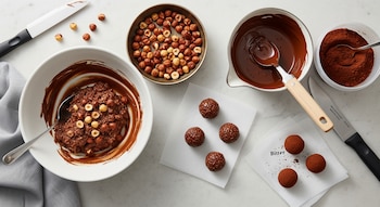 Masa de chocolate y avellanas en un bol, avellanas tostadas, chocolate fundido, cacao amargo, bombones con virutas sobre papel, bombones con cacao sobre papel, cuchillos.