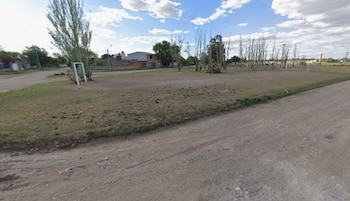 Un camino de tierra en primer plano, un terreno con un arco de fútbol, árboles y varias casas al fondo bajo un cielo con nubes